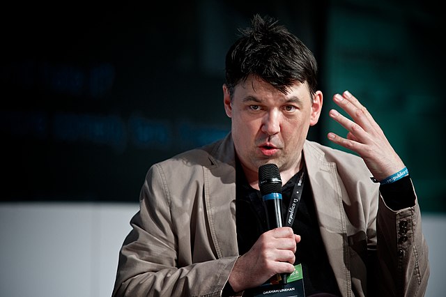 The picture shows him wearing a beige blazer jacket and holding a microphone. He’s giving a speech and gesturing towards his head with his off hand.