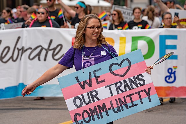 A photo of a woman at a Pride event holding a sign painted with the trans flag that reads "We heart our trans community".