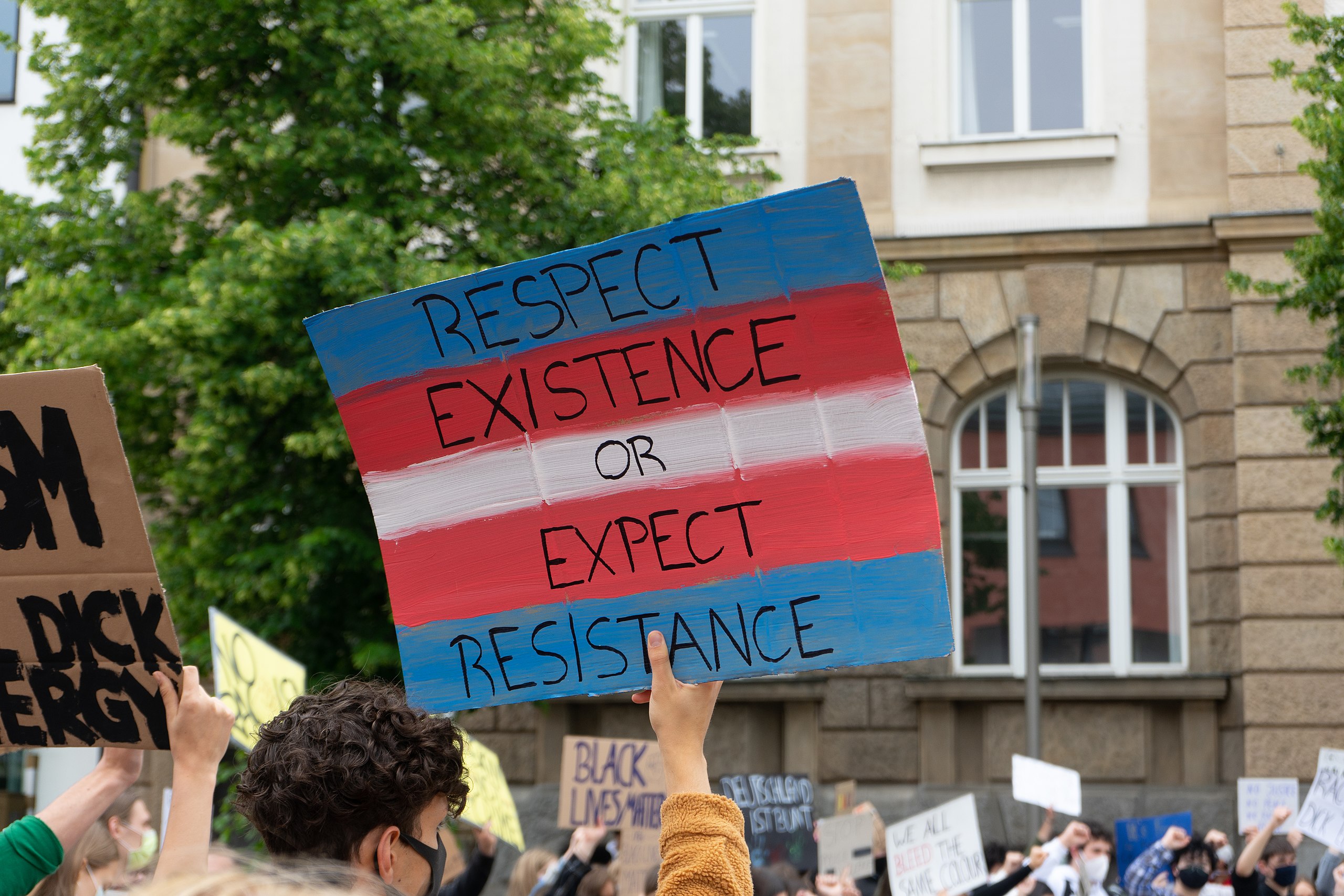 A photo from a black lives matter protest in Hof in 2020. Someone holds a piece of cardboard painted with a transflag reading "respect existence or...