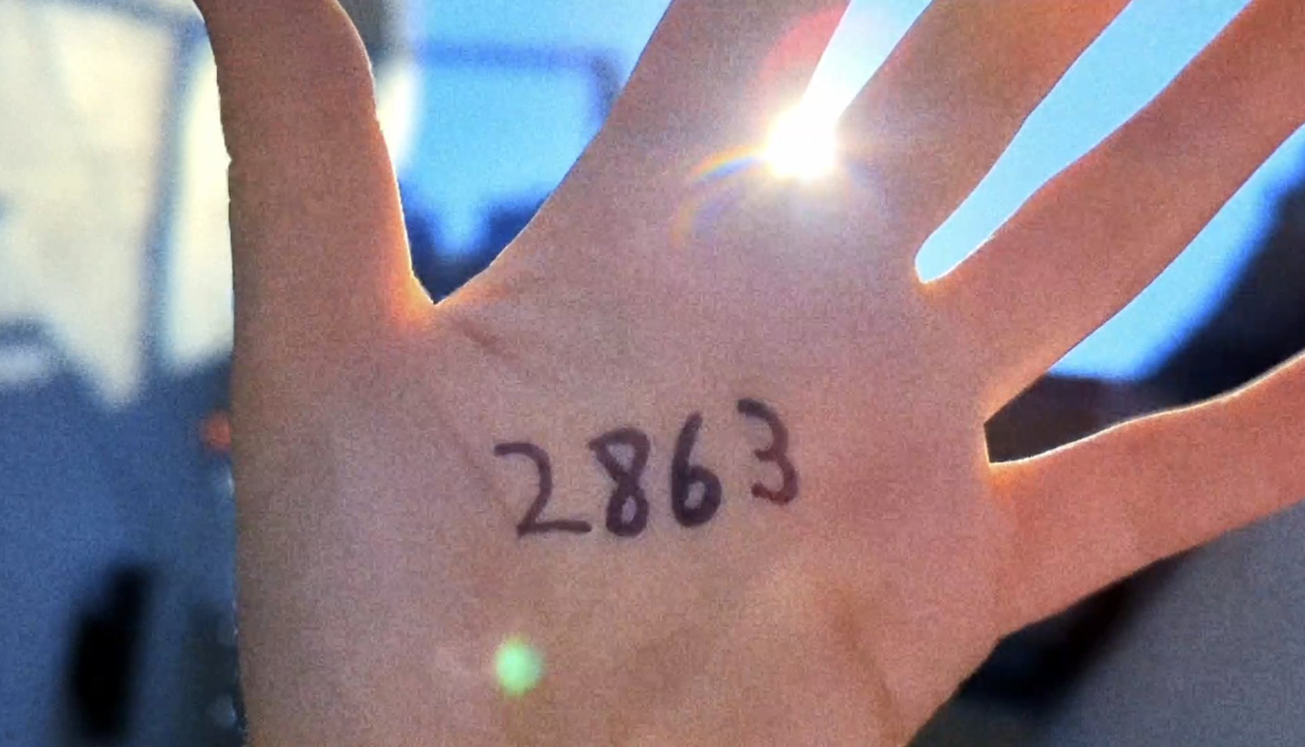 Promotional image for Again Again. The image shows a hand held up against the sun, written in the palm is the number "2863" - which represents the ...