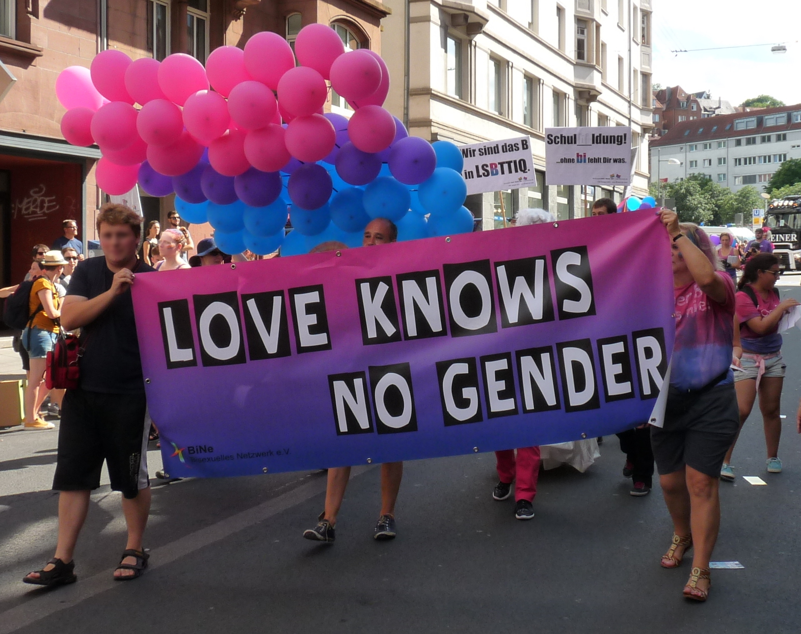 Bi visibility day: a group of people carry a large banner painted in the bisexual flag colours that reads "Love knows no gender" and a large bisexu...