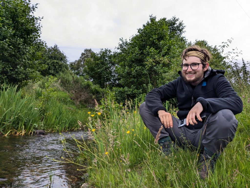A photo of Jay Hulme squatting near a river near the the Monadhliath Mountains