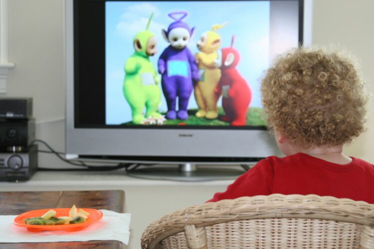 A toddler sits in front of a very 90s television watching Teletubbies. The toddler has very curly dark blonde hair, a red jumper and is sitting on ...