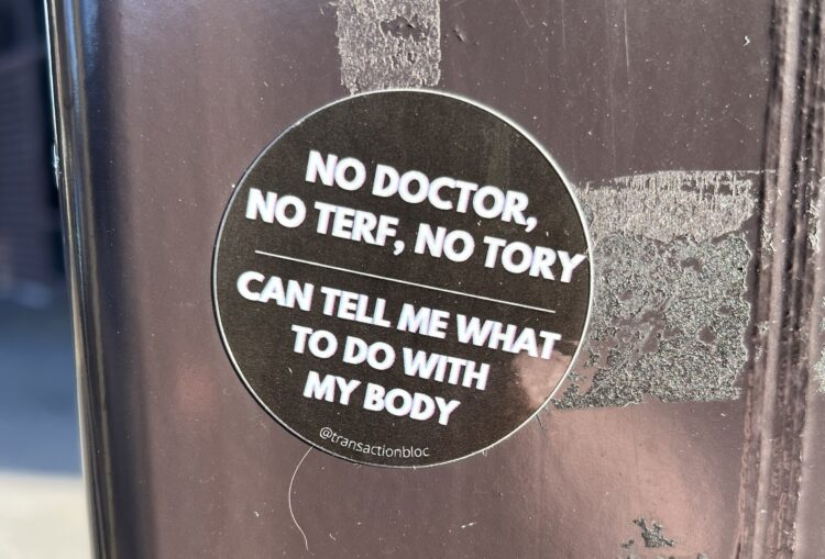 An anti-transphobia sticker stuck to a stainless steel surface out in the wild. It is a black circle with white outline and white text which reads ...