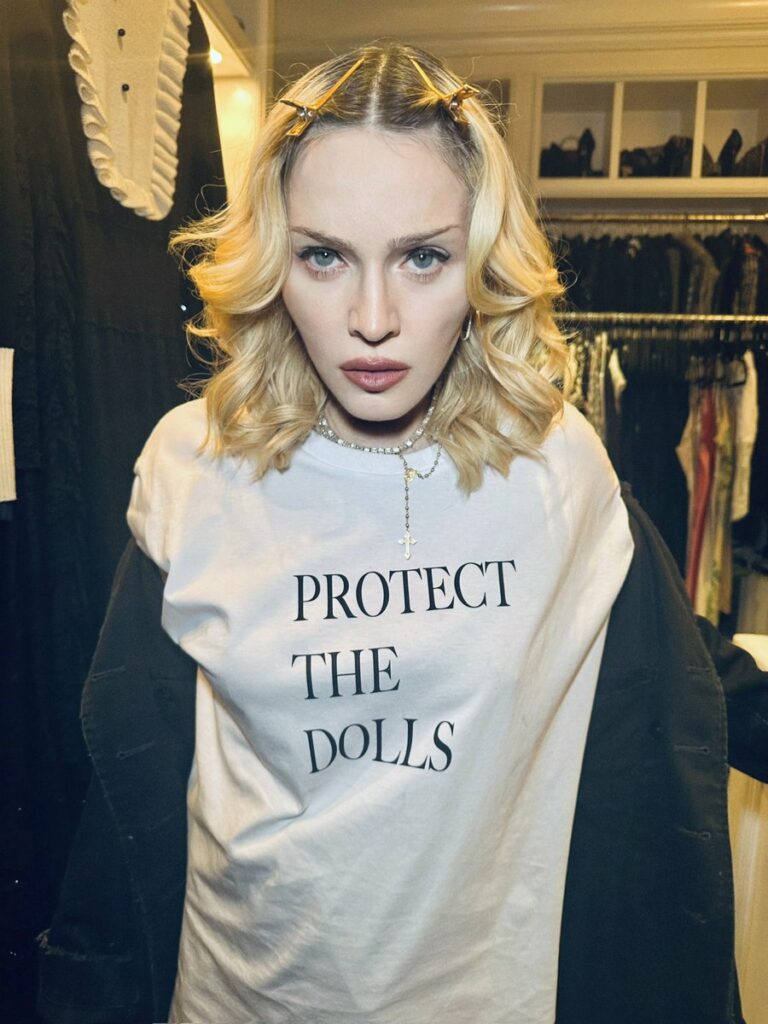 A photo of Madonna, presumably in a closet she owns as rails of clothes and shoes can be seen behind her. She is wearing a black jacket and a plain white t-shirt which reads "PROTECT THE DOLLS" in all capital letters.