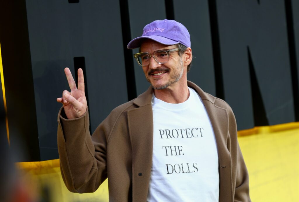 A photo of Pedro Pascal giving a peace sign to someone off camera at an event. He is wearing a light purple hat, brown coat and a plain white t-shirt which reads "PROTECT THE DOLLS" in all capital letters.