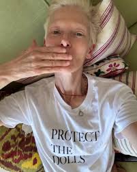 A photo of Tilda Swindon laying down on her back and blowing a kiss while showing off a plain white t-shirt that reads "PROTECT THE DOLLS" in all capital letters.