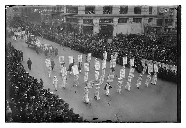 An image of a suffragettes march in New York dated October 1915. There are hundreds if not thousands of people watching as about 20 women march with signs I can't read because the picture is old and small and I have bad eyes.