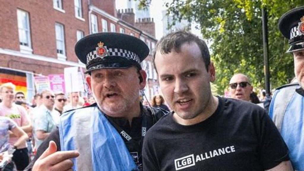 A photo of LGB Alliance supporter Alex Bramham being escorted away from Manchester Pride by Police Liason Officers. He is notably not wearing a hat.