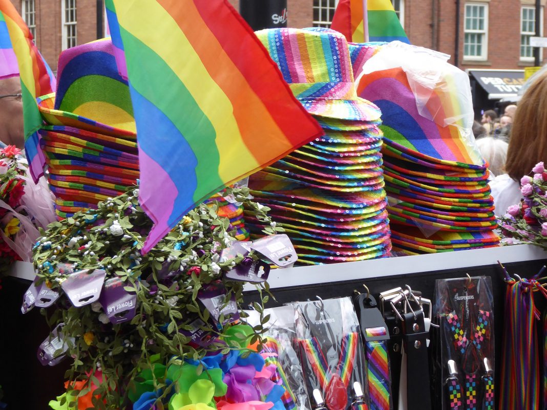 A rainbow merchandise stand at Manchester Pride, August 2016. It shows various rainbow themed lanyards and hats all piled up next to a small pride flag.