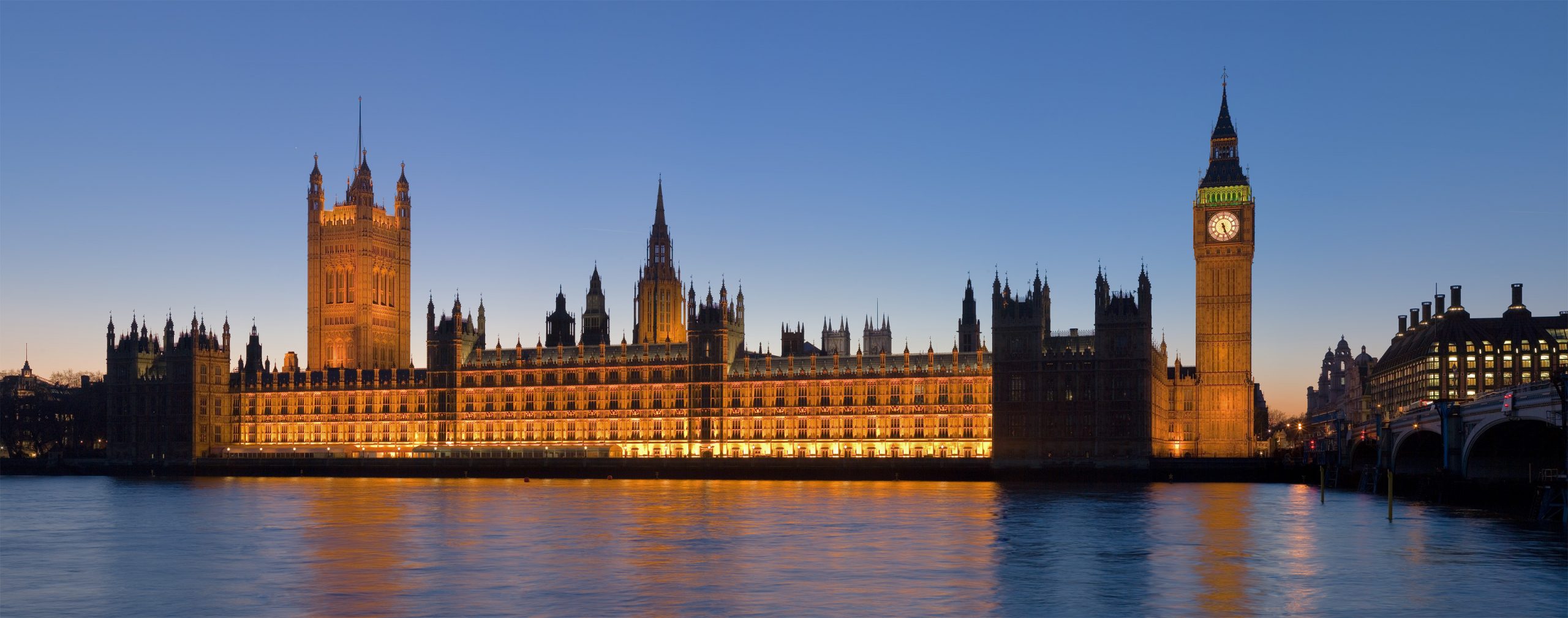 A photo of the Palace of Westminster taken from across the river during the evening. There's a real beautiful contrast between the fiery oranges lighting up the walls of the palace and the peaceful blue sky above.
