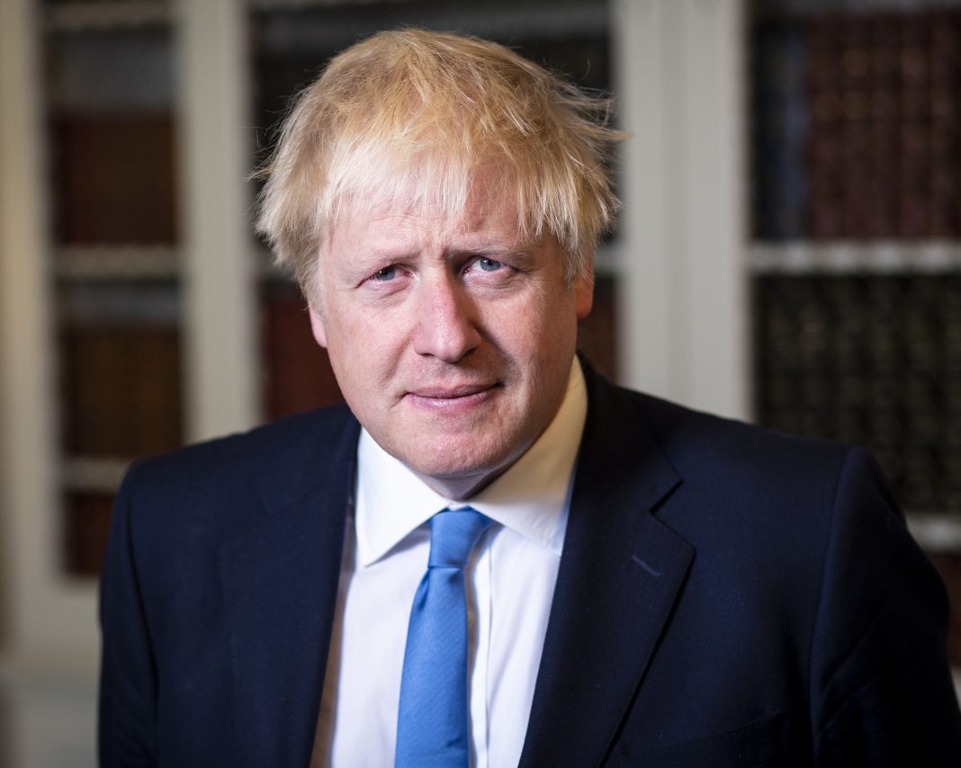 A portrait of Boris Johnson wearing a blue tie, navy blue suit and a blurred out book case behind him.
