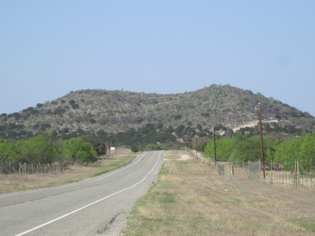 A photo of Highway 55 into Uvalde County, Texas. It simply shows a road heading towards a mountain in the distance, with lots of short bushy trees on either side.