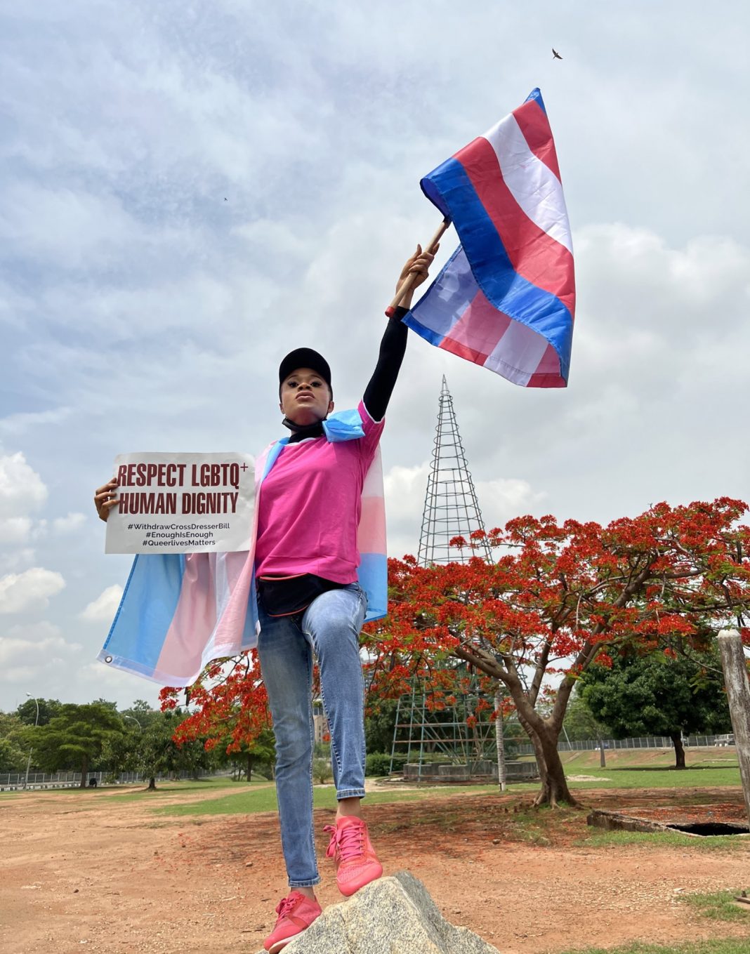 A Nigerian protestor stands on top of a rock triumphantly holding a trans flag high. In her other hand she holds a sign reading 