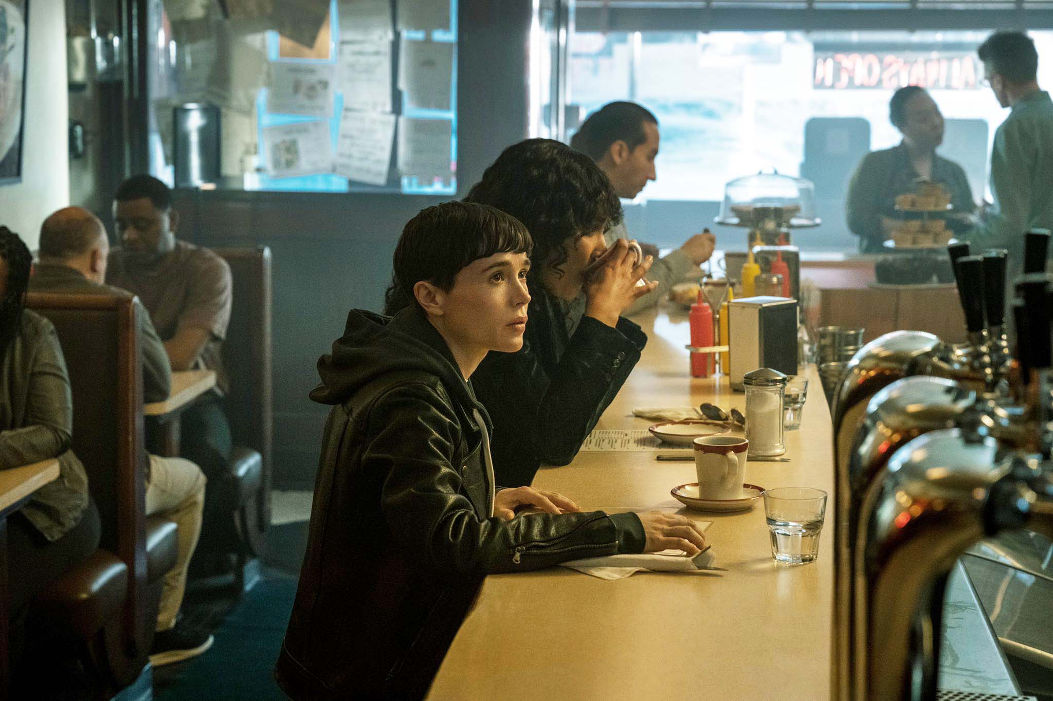 A still from The Umbrella Academy Season 3 showing Viktor Hargeeves (Elliot Page) sitting at a crowded bar