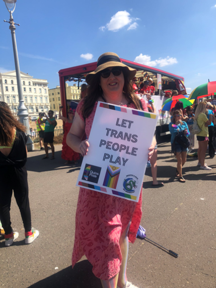 A person in a long pink dress wearing a straw hat and sunglasses holds a sign that says 'LET TRANS PEOPLE PLAY'