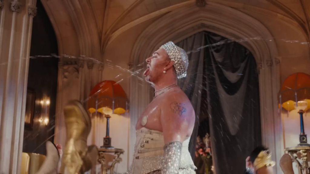 Sam Smith wearing a corset, nipple pasties a bejewelled sailor hat and long shiny gloves holding their tongue out while being sprayed with water in a fancy house surrounded by queers